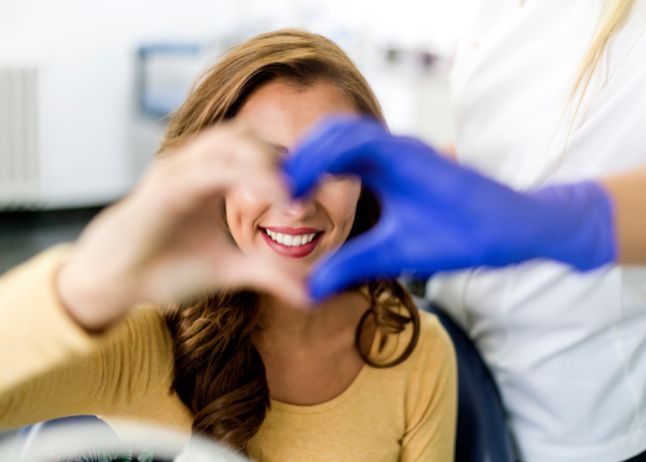 Woman smiles in dental office, hands form heart over her face. Blue-gloved hands in foreground.