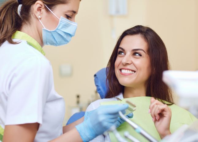 Dentist with mask and gloves examining a smiling woman in a dental chair.
