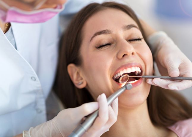 Woman at dentist, mouth open, being examined. Dentist in pink mask and white coat, tools visible.