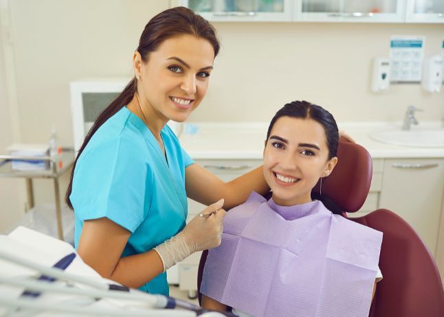 Dentist in blue scrubs with a patient in a dental chair smiling in a bright dental office.
