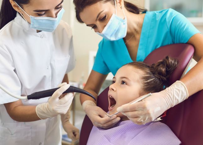 Dentists examining a young girl's teeth. Two adults in masks and gloves. Girl  in dentist chair, mouth open.