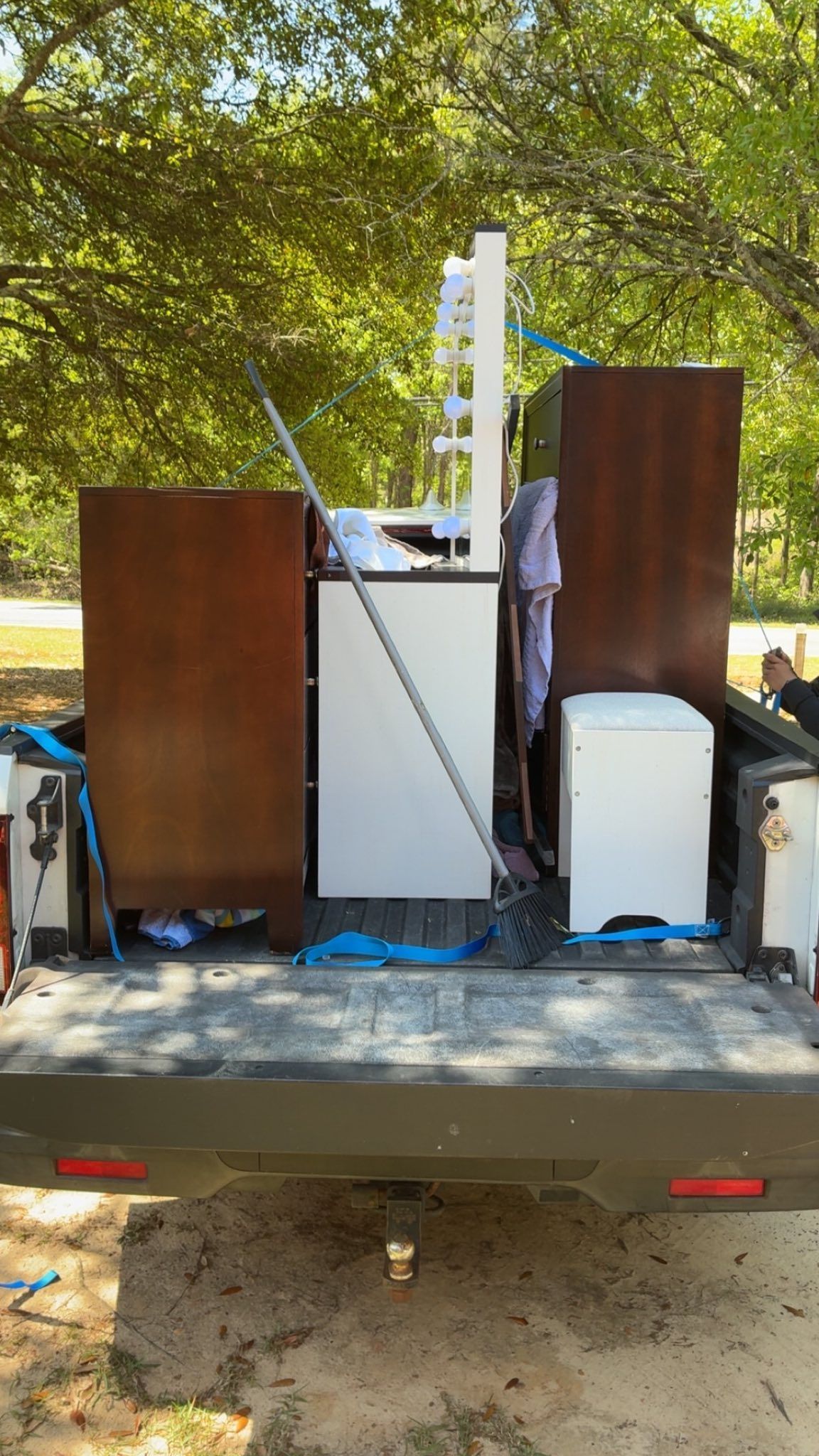 Furniture, including wooden cabinets and a white shelf unit, secured with blue straps in the bed of a pickup truck.