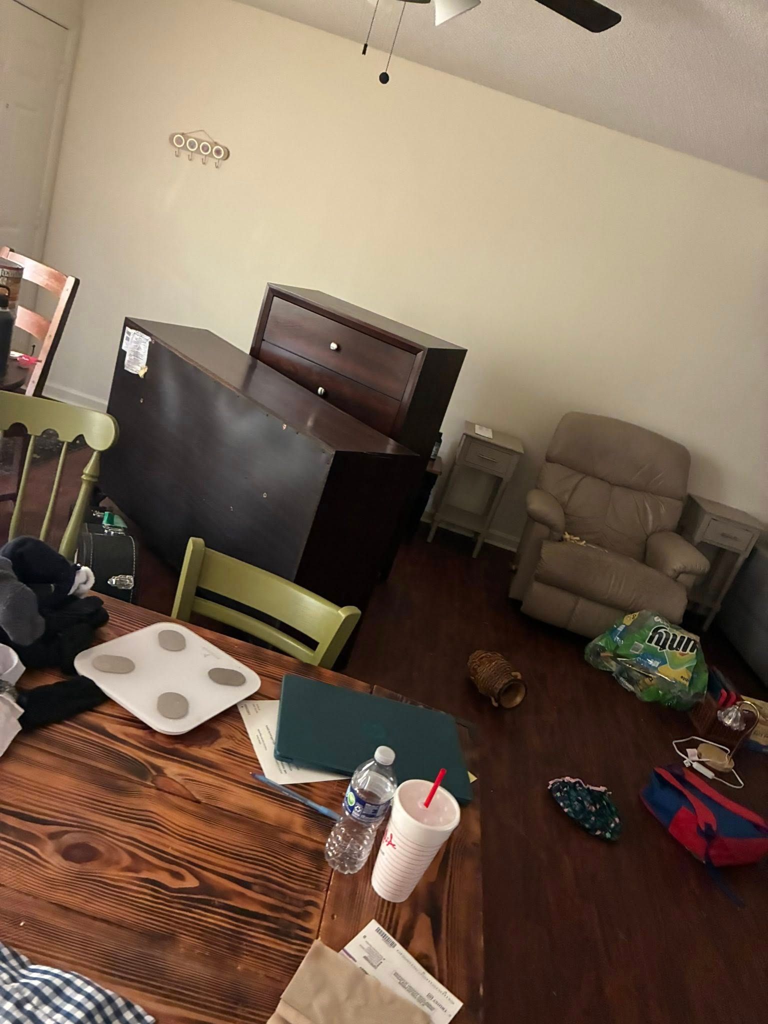 A dark wooden dresser sits on a dark floor in a room with a light-colored armchair, a wooden table, and scattered items.