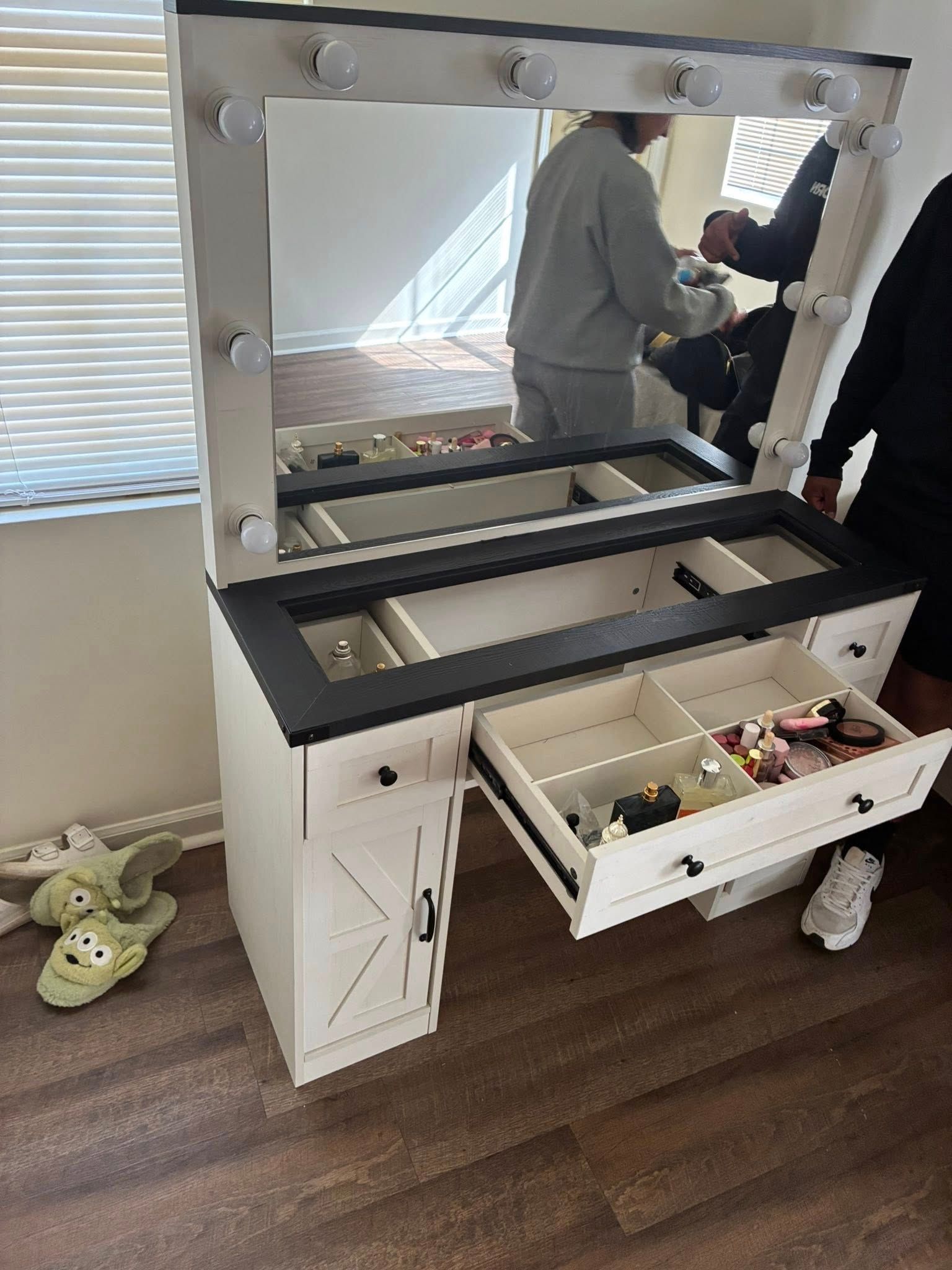A white makeup vanity with black accents, a lighted mirror, and an open drawer, positioned in a room with hardwood floors.