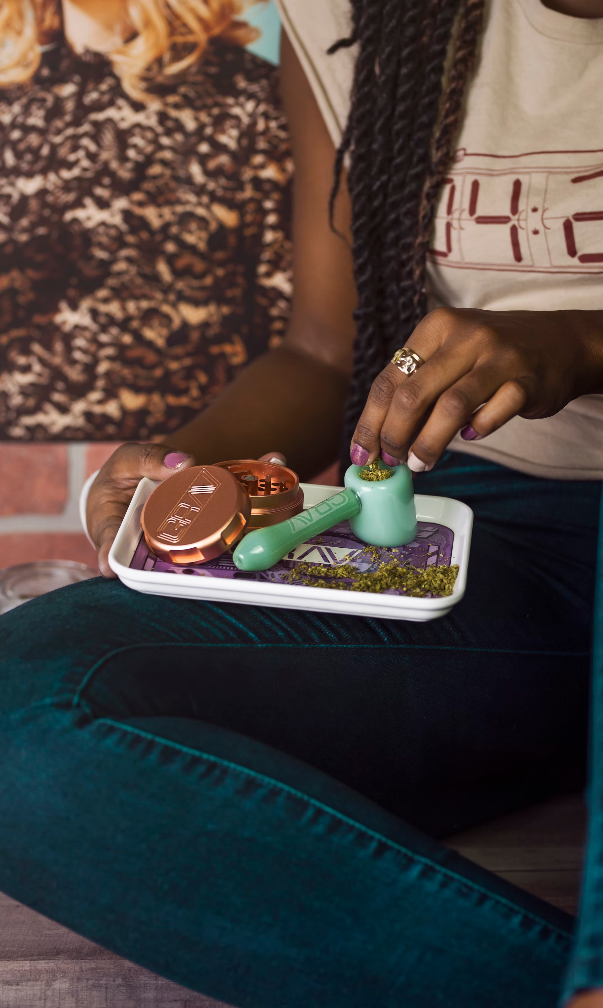 Person holding a tray with a bong, weed, and accessories. The person is seated and wearing teal jeans.