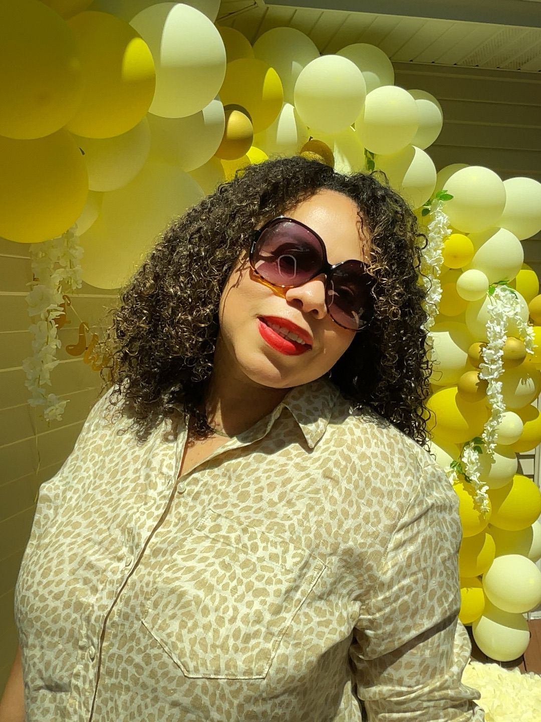 A woman wearing sunglasses and a leopard print shirt is standing in front of balloons.