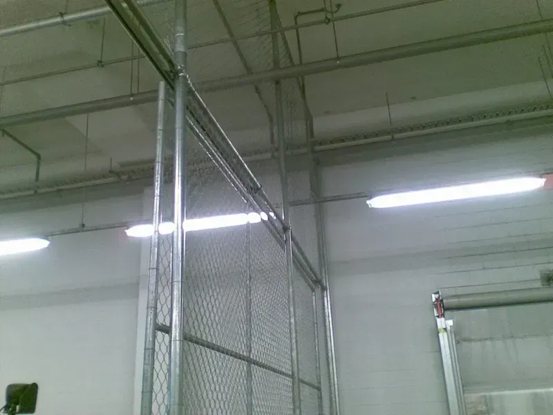 A chain-link fence partition inside a warehouse with overhead industrial lighting and exposed pipes on the ceiling.