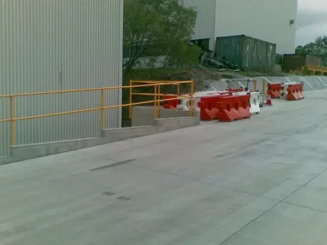 A concrete loading ramp with yellow metal railings alongside orange and white plastic safety barriers on a paved lot.
