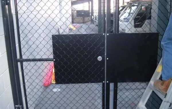 A black security plate installed over a chain-link fence gate, with a person standing on a ladder to the right.