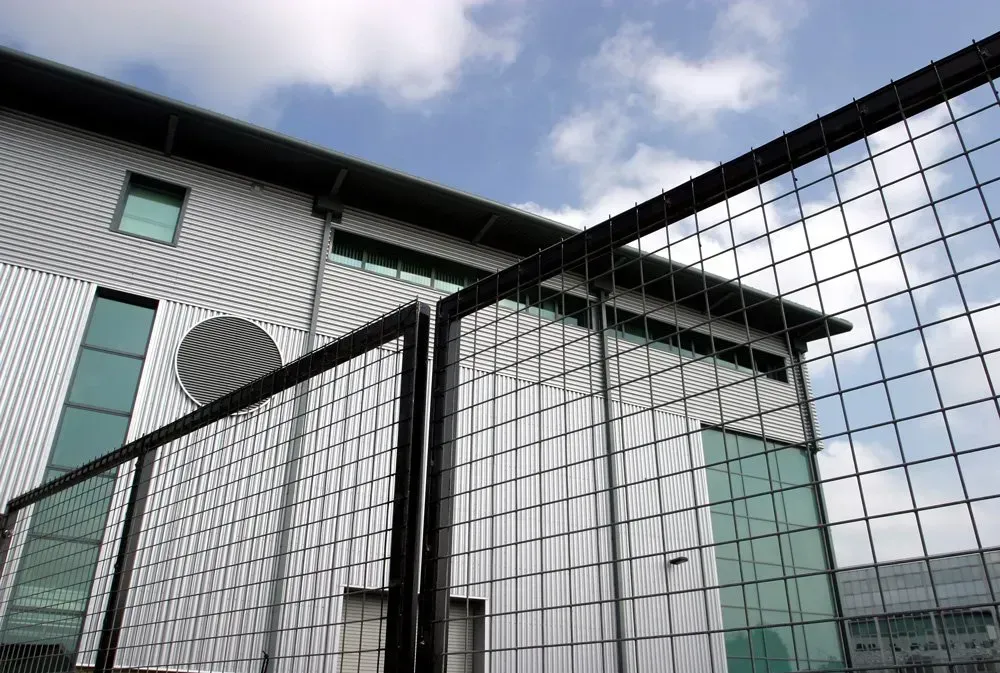 A modern industrial-style building sits behind a high wire-mesh fence topped with barbed wire against a cloudy sky.