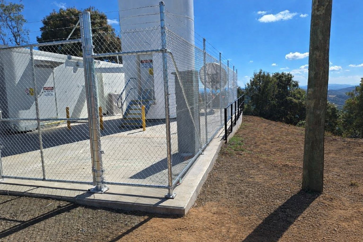 A fenced utility site with a white tower and equipment enclosure, situated on a grassy hill under a bright blue sky.