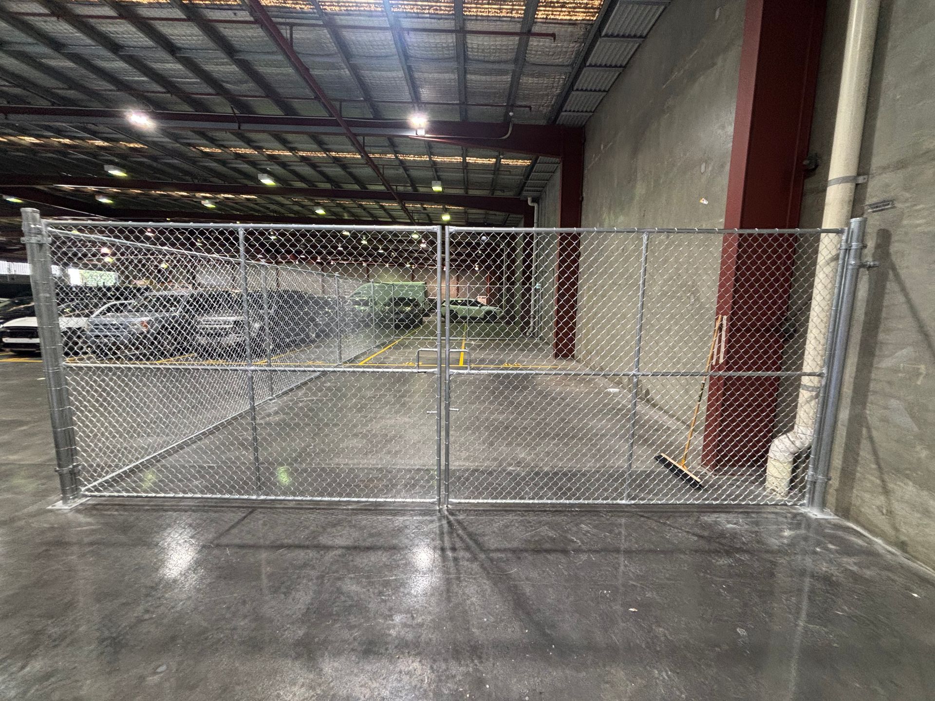 A metal chain-link gate separates a warehouse floor from a parking area containing cars.