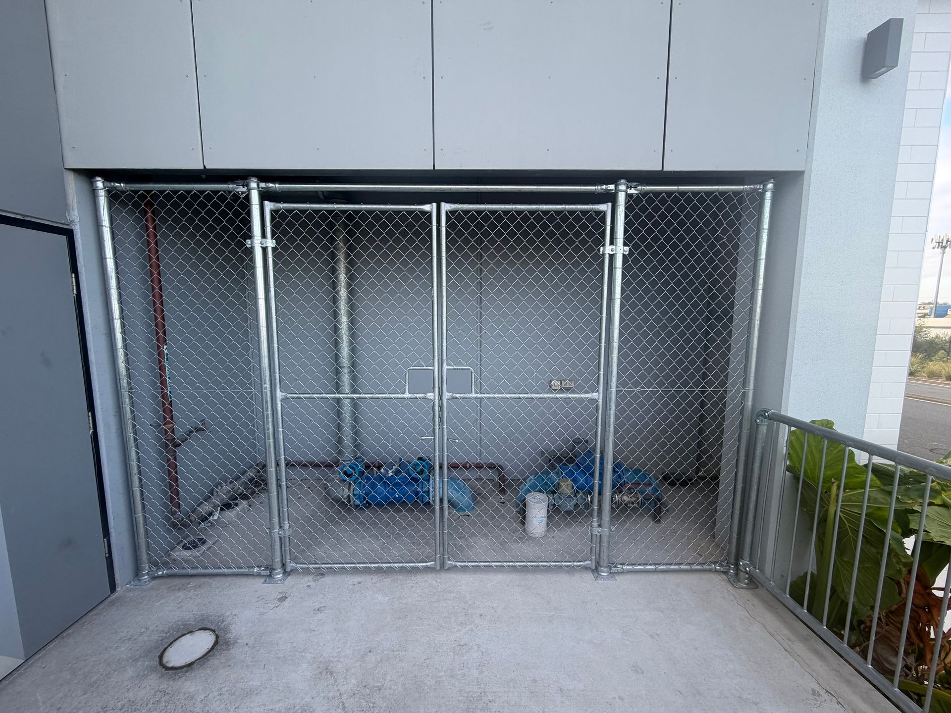 A chain-link fence enclosure with double doors protects two blue pumps and piping against a building wall.