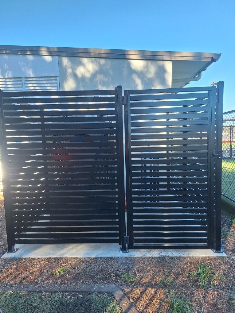 A black horizontal slat privacy screen and gate installed on a concrete pad against the side of a building.
