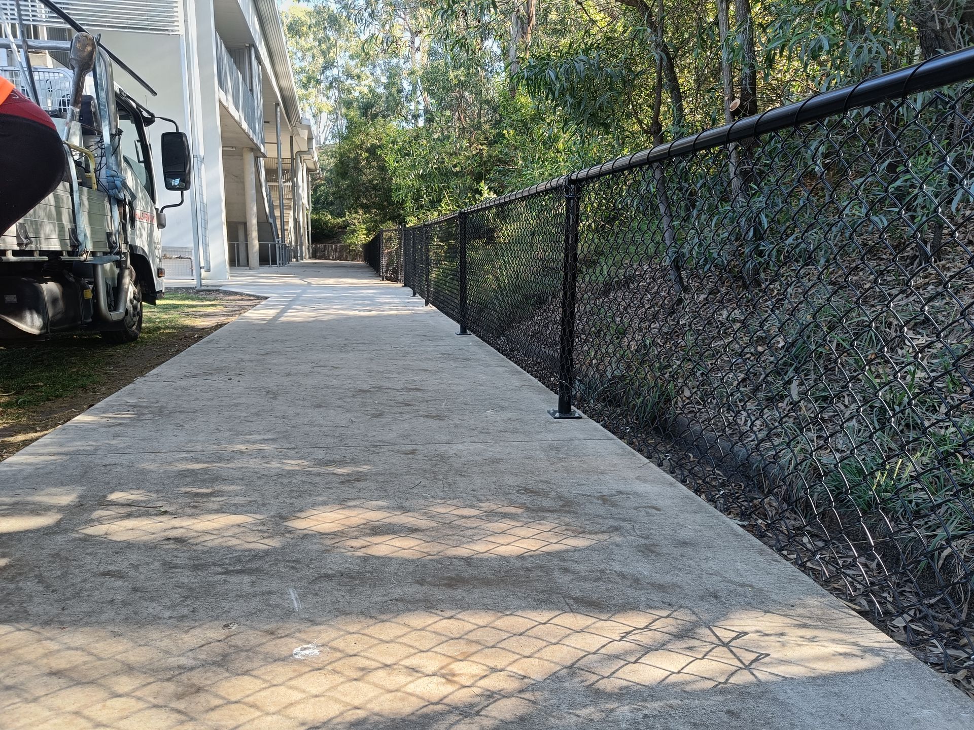 A paved sidewalk runs alongside a black chain-link fence bordering a wooded area next to a building.