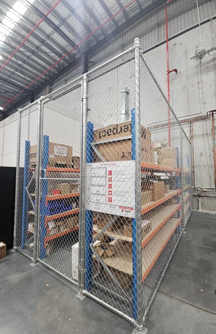 Industrial warehouse cage with chain-link fencing enclosing blue metal shelving units filled with various boxed items.