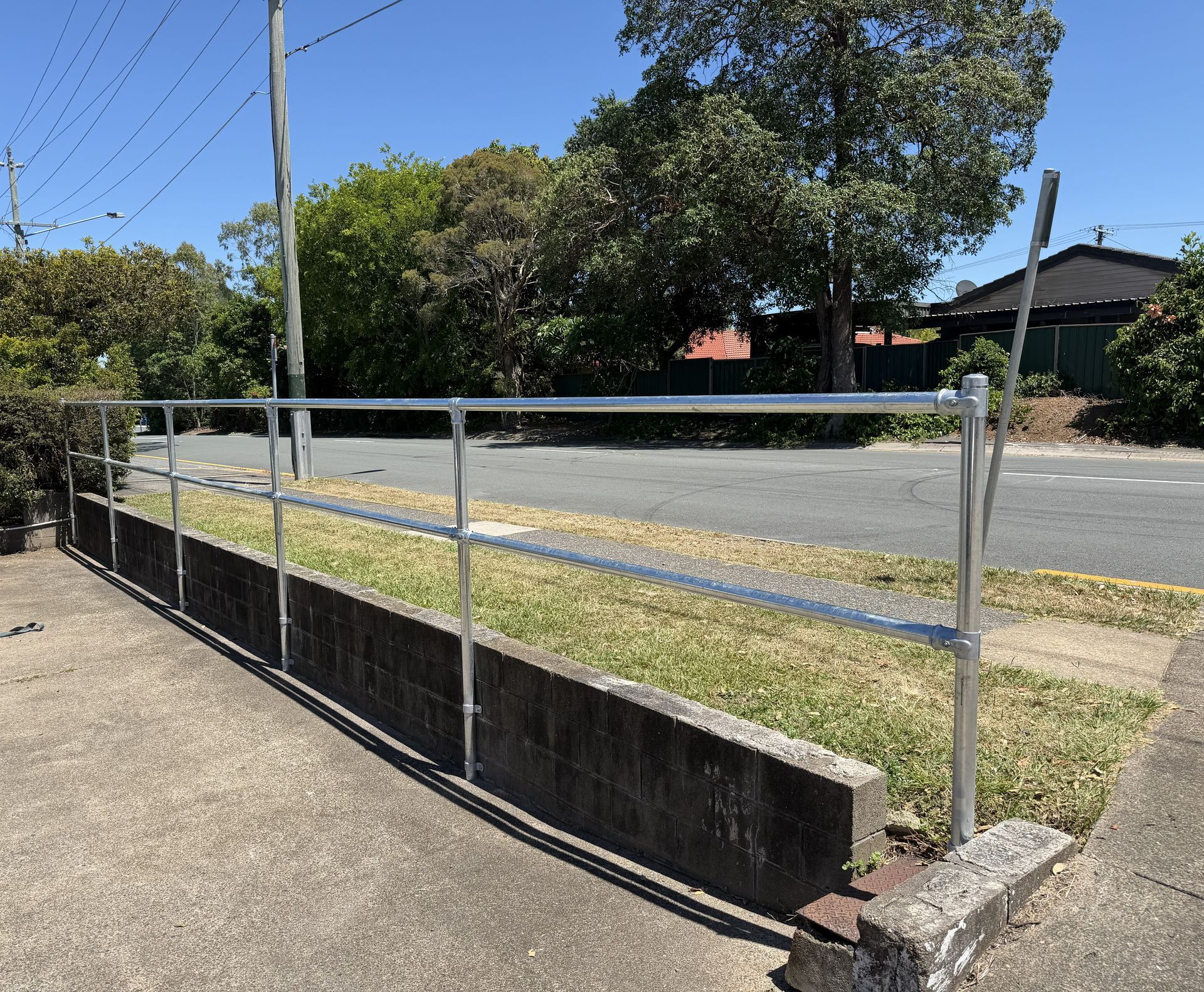 A silver metal safety railing stands on a concrete retaining wall alongside a grassy verge and a residential street.