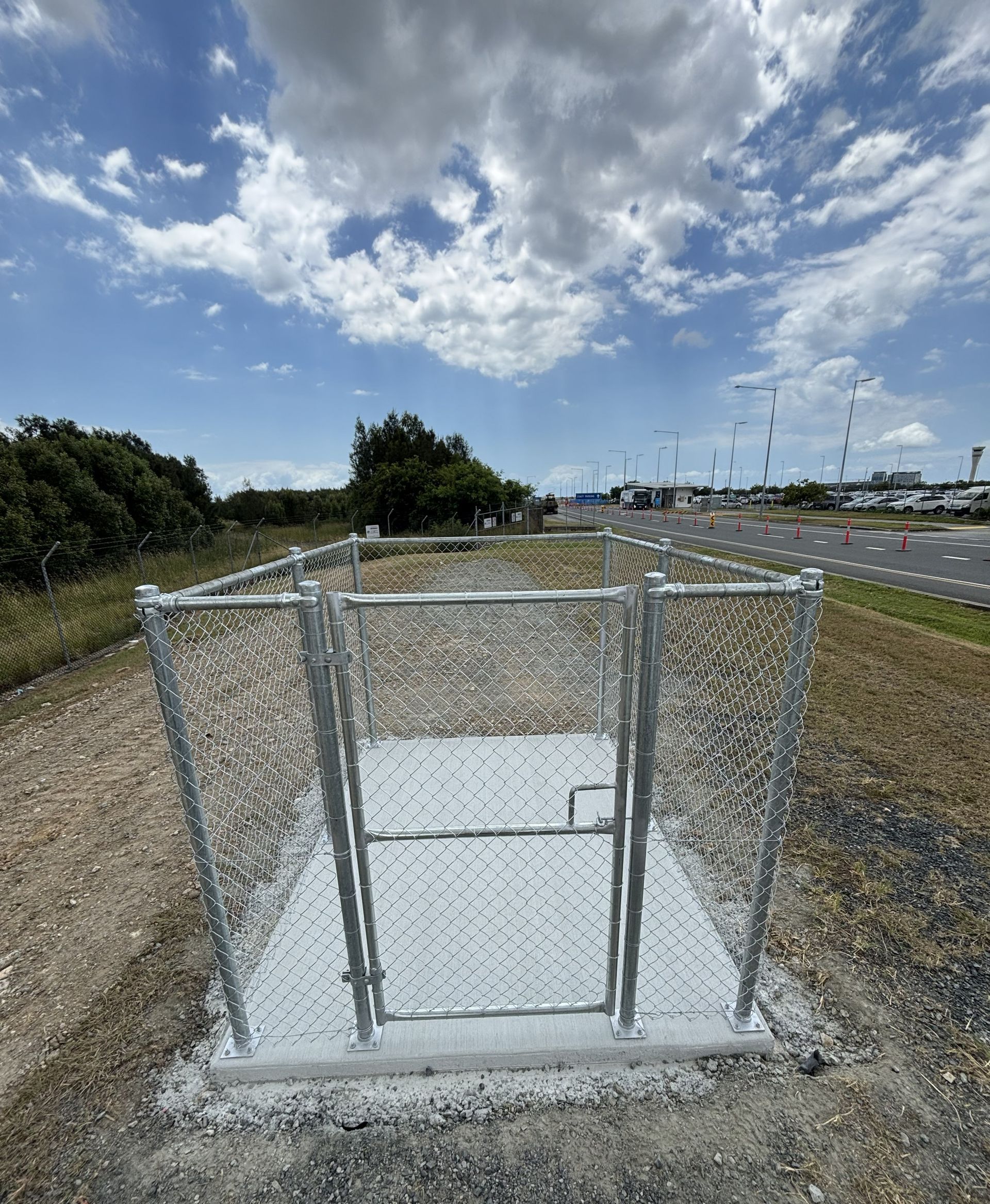 A chain-link fenced enclosure with a metal gate sits on a concrete pad surrounded by gravel under a blue, cloudy sky.