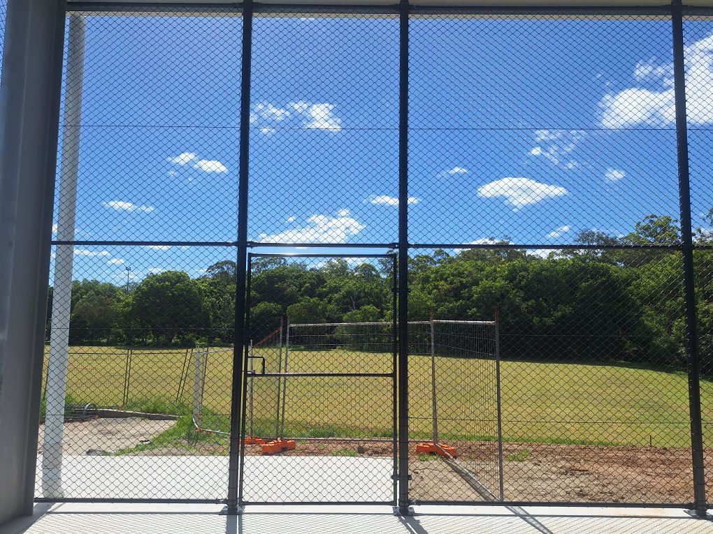 View through a chain-link fence of a grassy field, trees, and a bright blue sky with clouds.