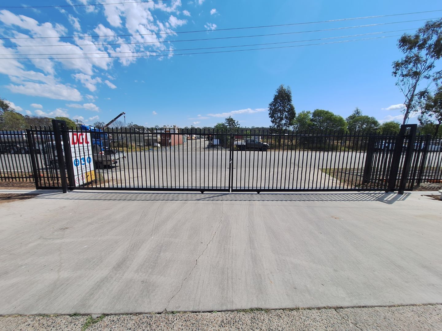 A wide black metal sliding gate blocks a paved entrance leading to a gravel yard with trees under a bright blue sky.