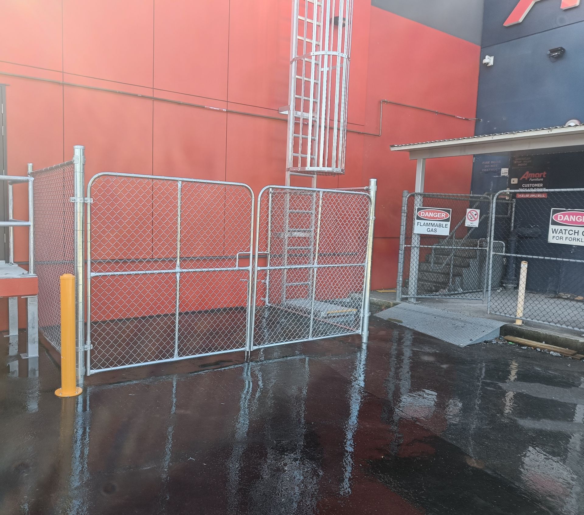 Chain-link fenced enclosures and a vertical ladder mounted on a red building wall, with a small ramped area to the right.