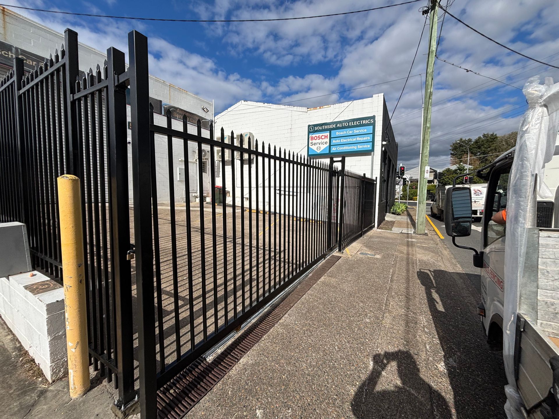 A black metal fence lines a sidewalk in front of a white building with a blue sign under a blue sky.