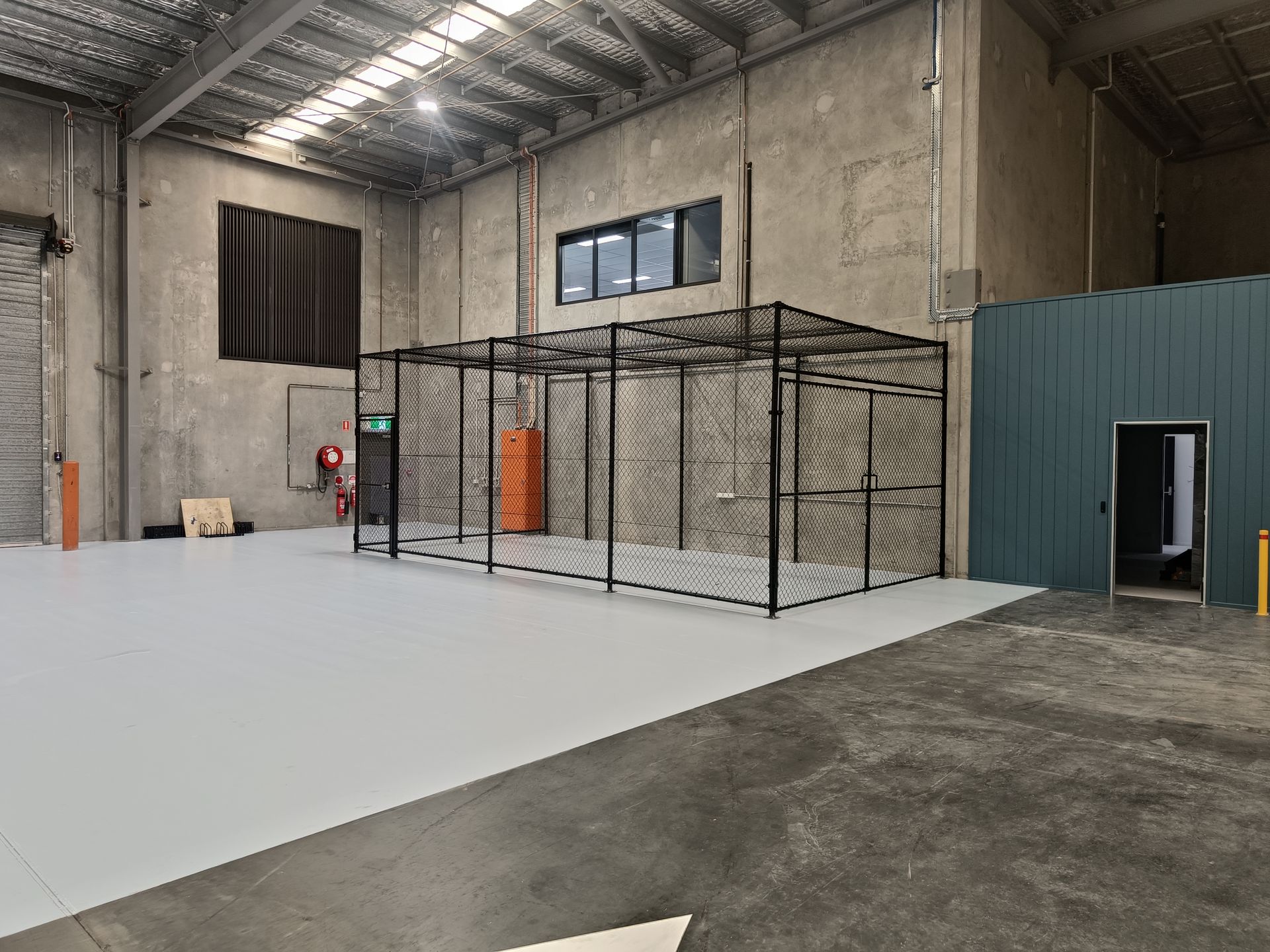 A large, empty warehouse interior featuring a high-ceiling space with a black chain-link enclosure set on a gray floor.