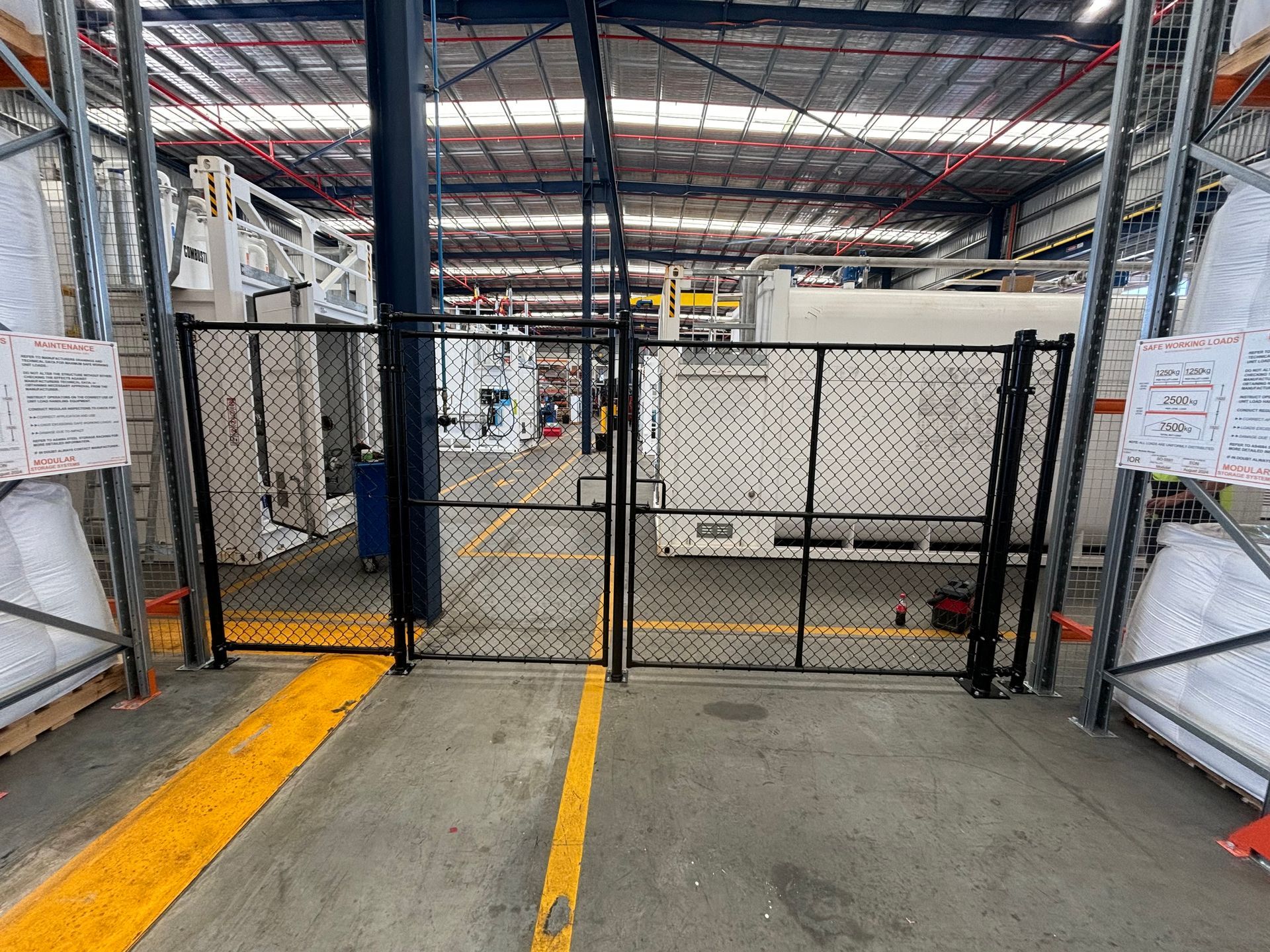 A fenced-off warehouse interior with yellow painted floor lines and large industrial containers in the background.