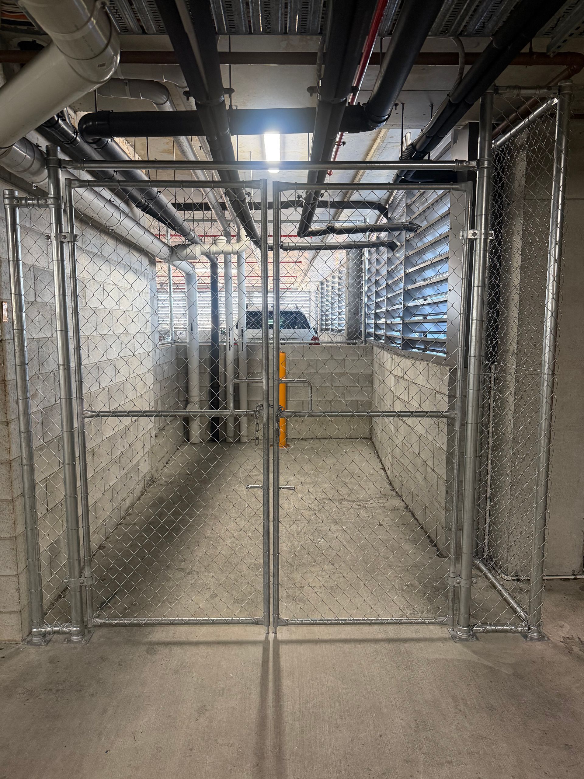 A chain-link storage cage with double gates inside a concrete underground parking garage with overhead utility pipes.