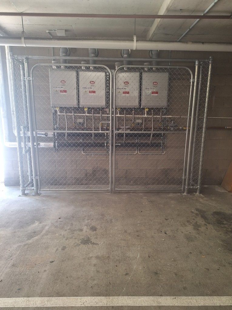 Four wall-mounted water heaters enclosed behind a wire chain-link fence in a parking garage.