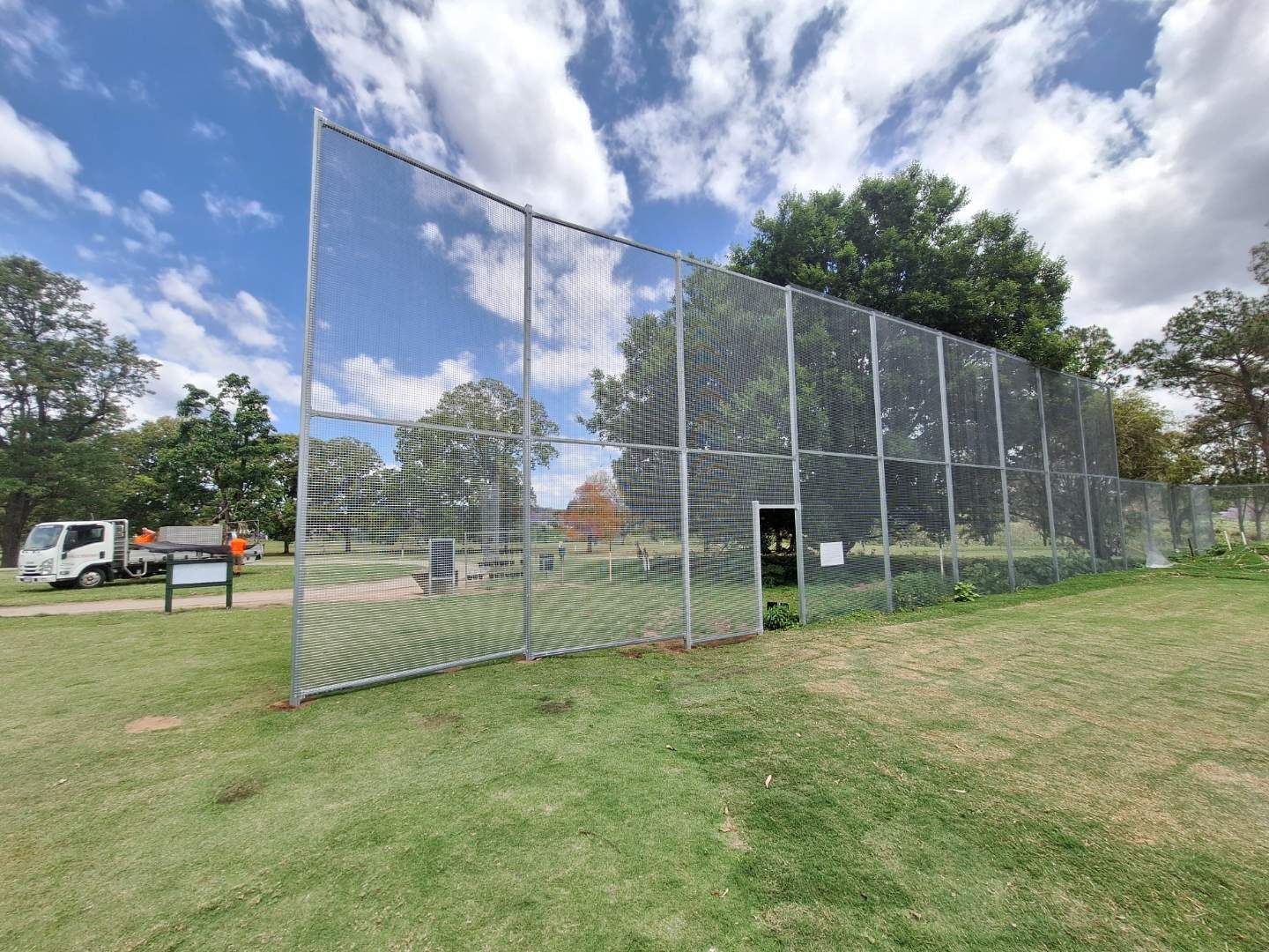 A tall metal chain-link fence stands in a grassy park field under a cloudy sky, with a white utility truck parked nearby.