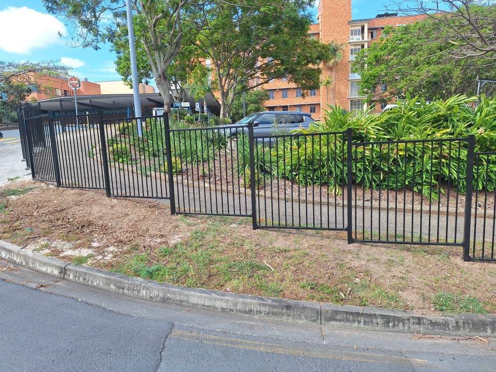 A black metal fence runs along a curb next to a green lawn, shrubs, and a brick apartment building in the background.