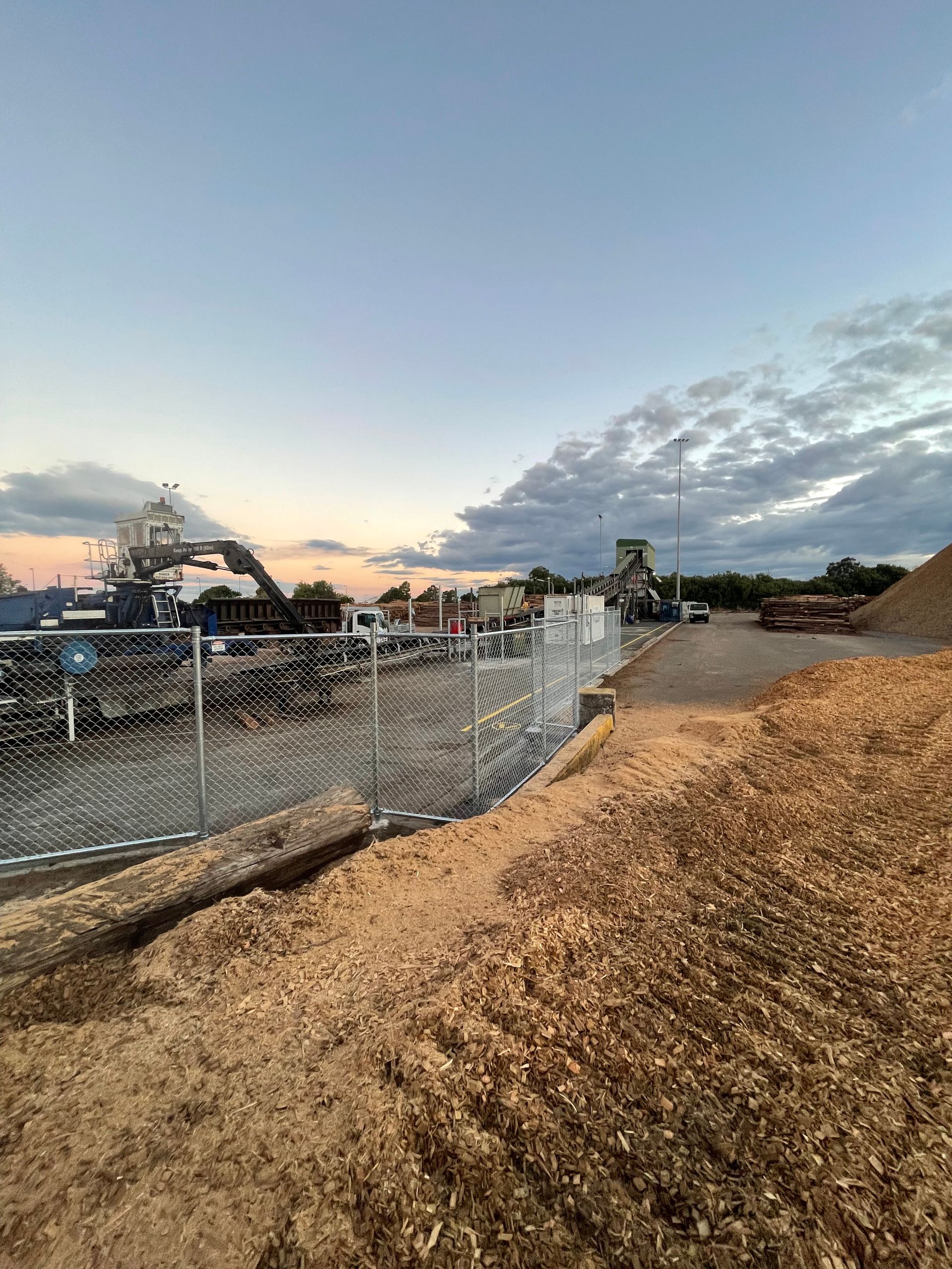 A fenced outdoor industrial site with a large pile of wood chips in the foreground under a cloudy sky at dusk.