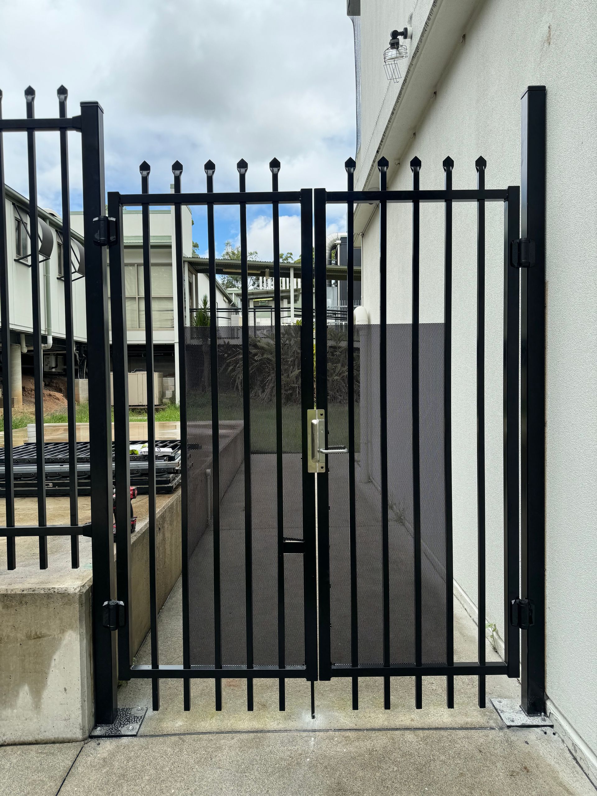 Black metal double gate with vertical bars and finial tops, installed on a concrete path against a white building wall.