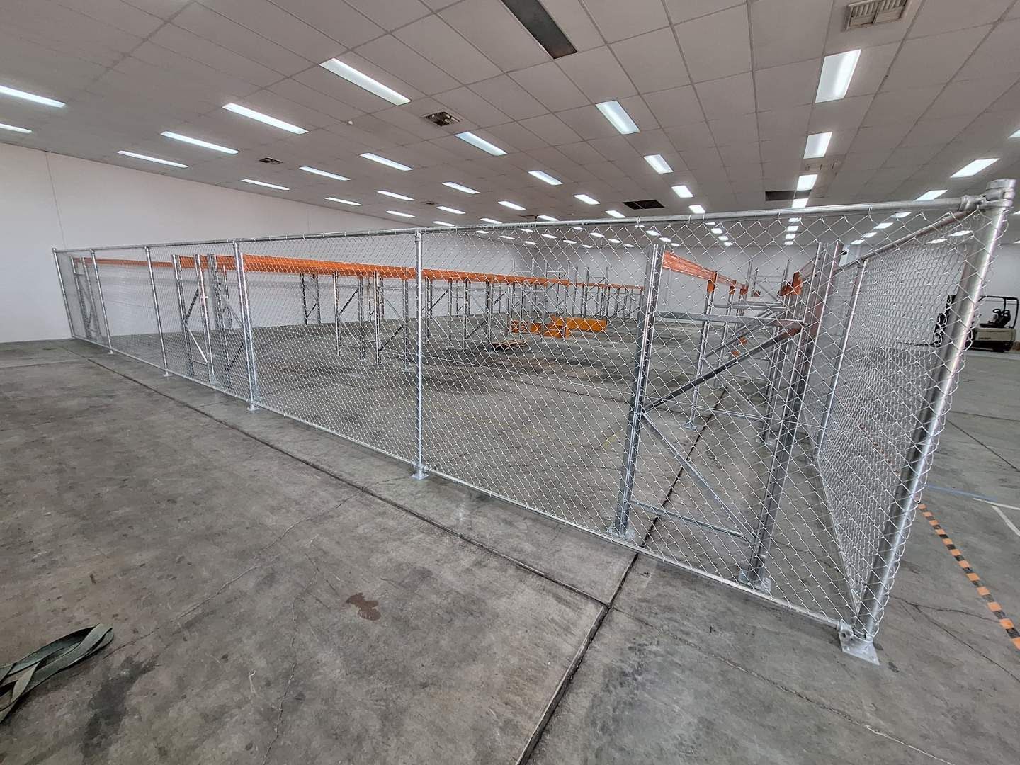 A chain-link fence enclosure stands in a brightly lit industrial warehouse, surrounding orange shelving units.