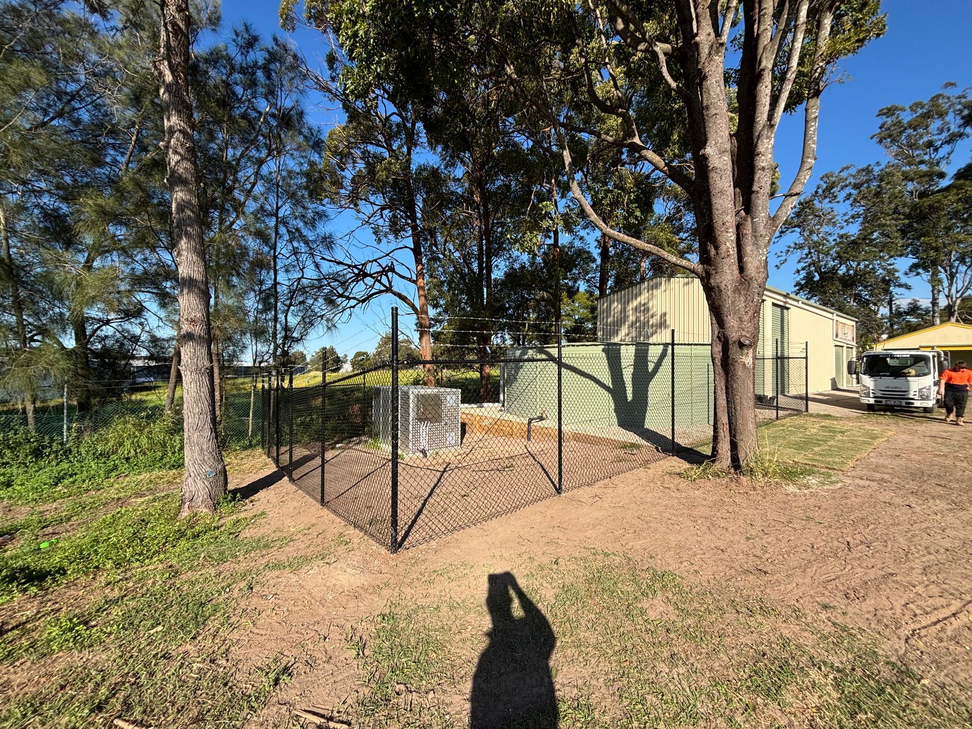 A fenced electrical equipment enclosure next to a large green water tank and a metal shed in a grassy, tree-filled area.