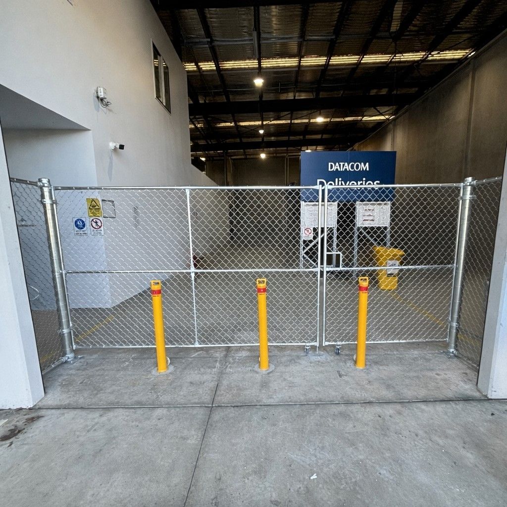 A wire fence separates a green grass sports field from a grassy path next to a building, with trees in the background.