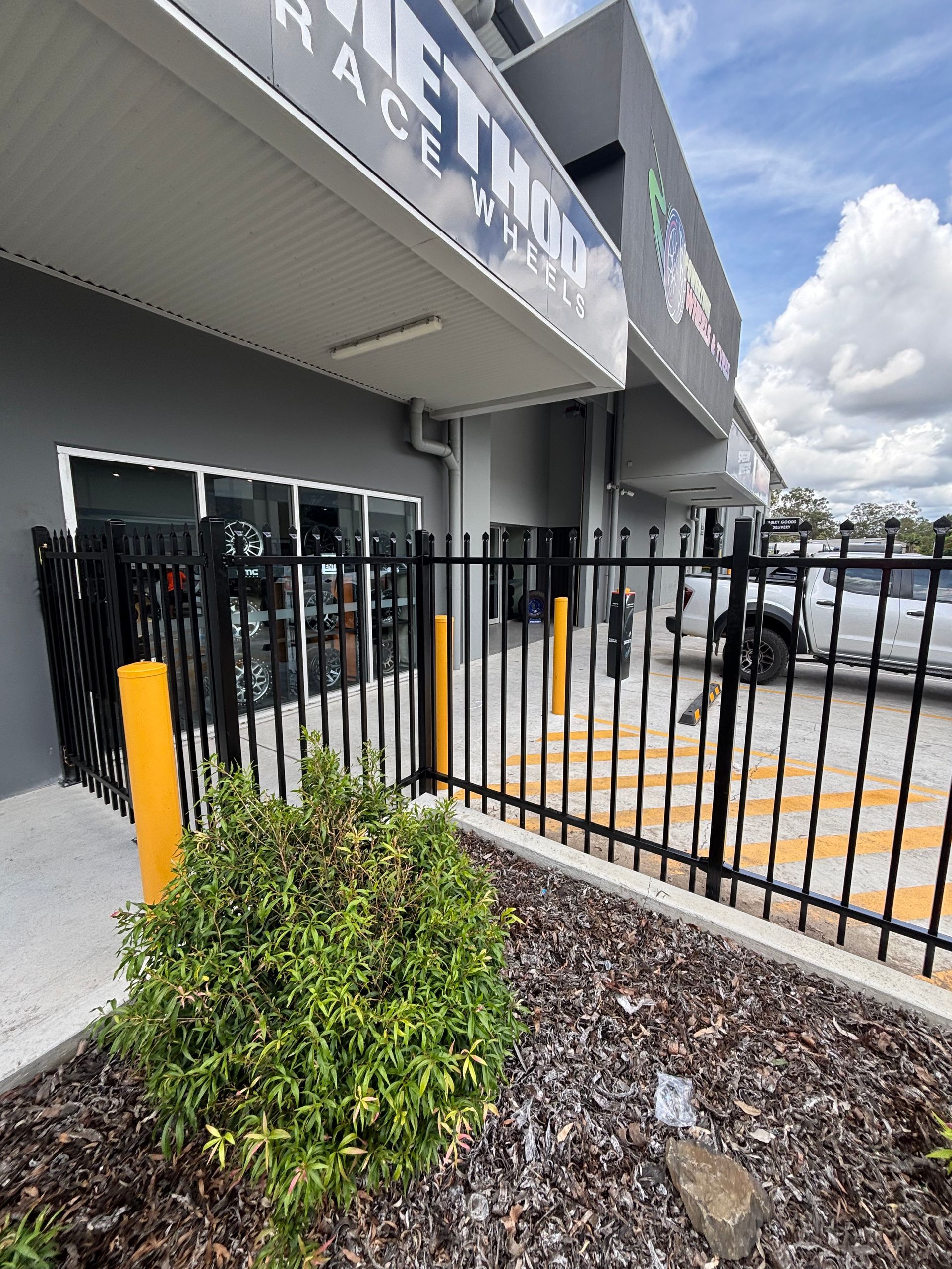 A storefront with a black metal fence and yellow bollards in front, under a blue sky.