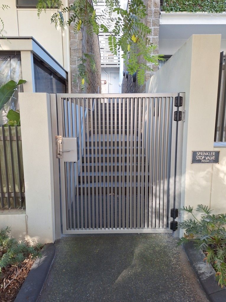 A silver-colored metal pedestrian gate featuring vertical slats, positioned between two light-colored concrete walls.