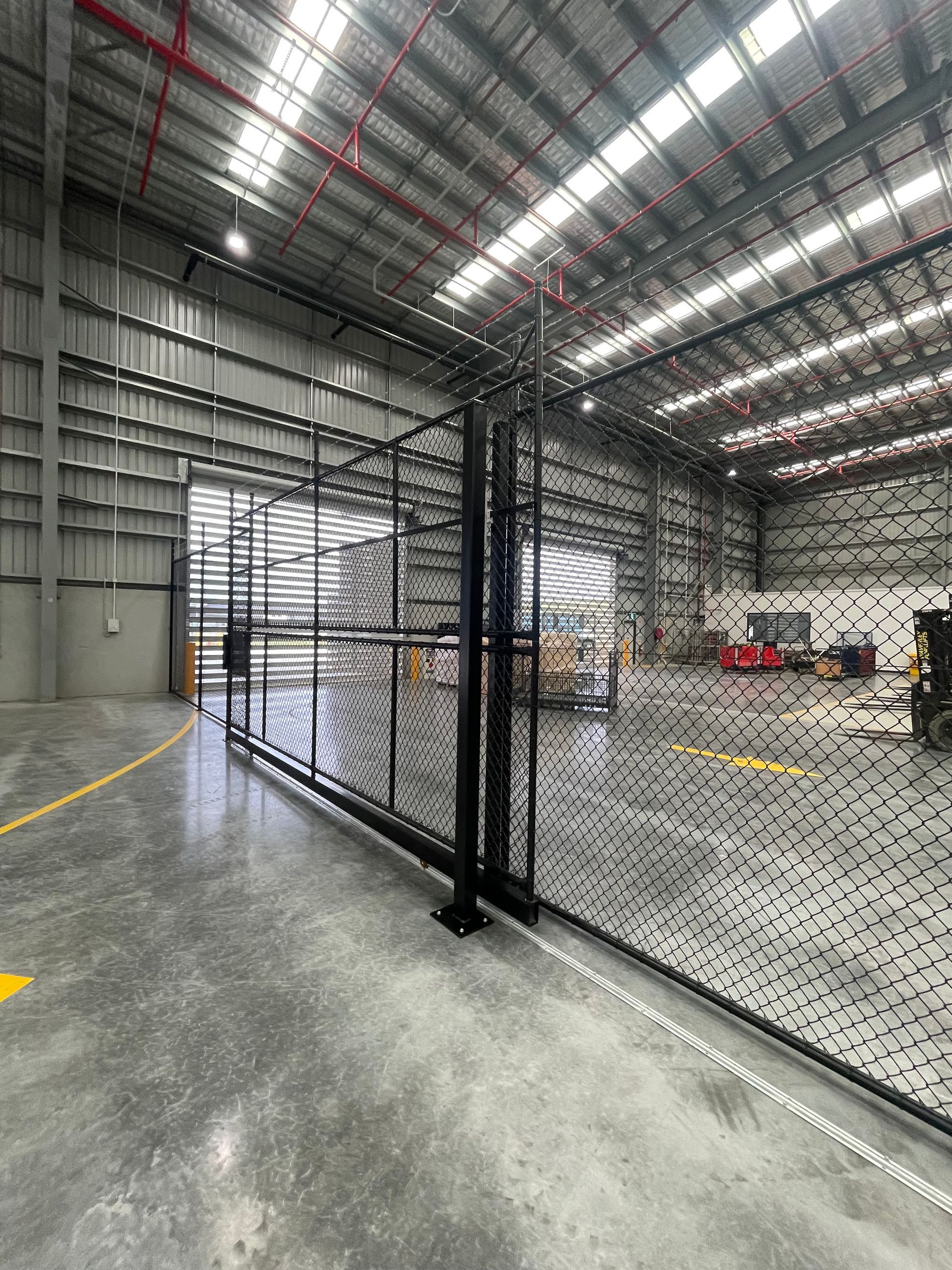 A black metal mesh security fence divides a spacious, brightly lit warehouse with a concrete floor and high ceilings.