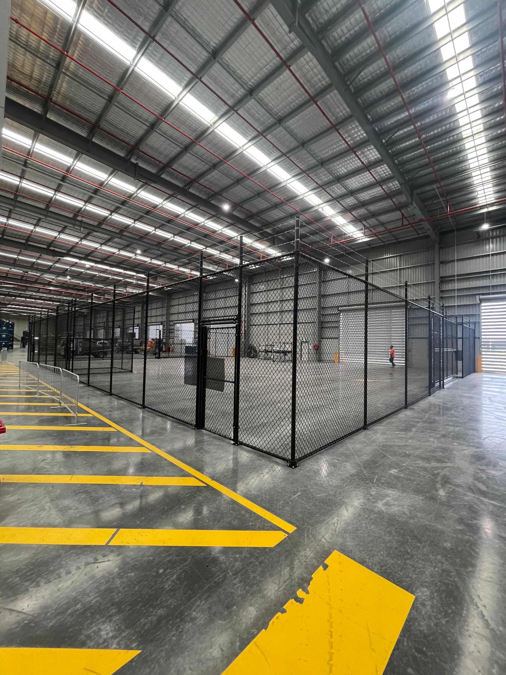 A chain-link fence partition inside a warehouse with overhead industrial lighting and exposed pipes on the ceiling.