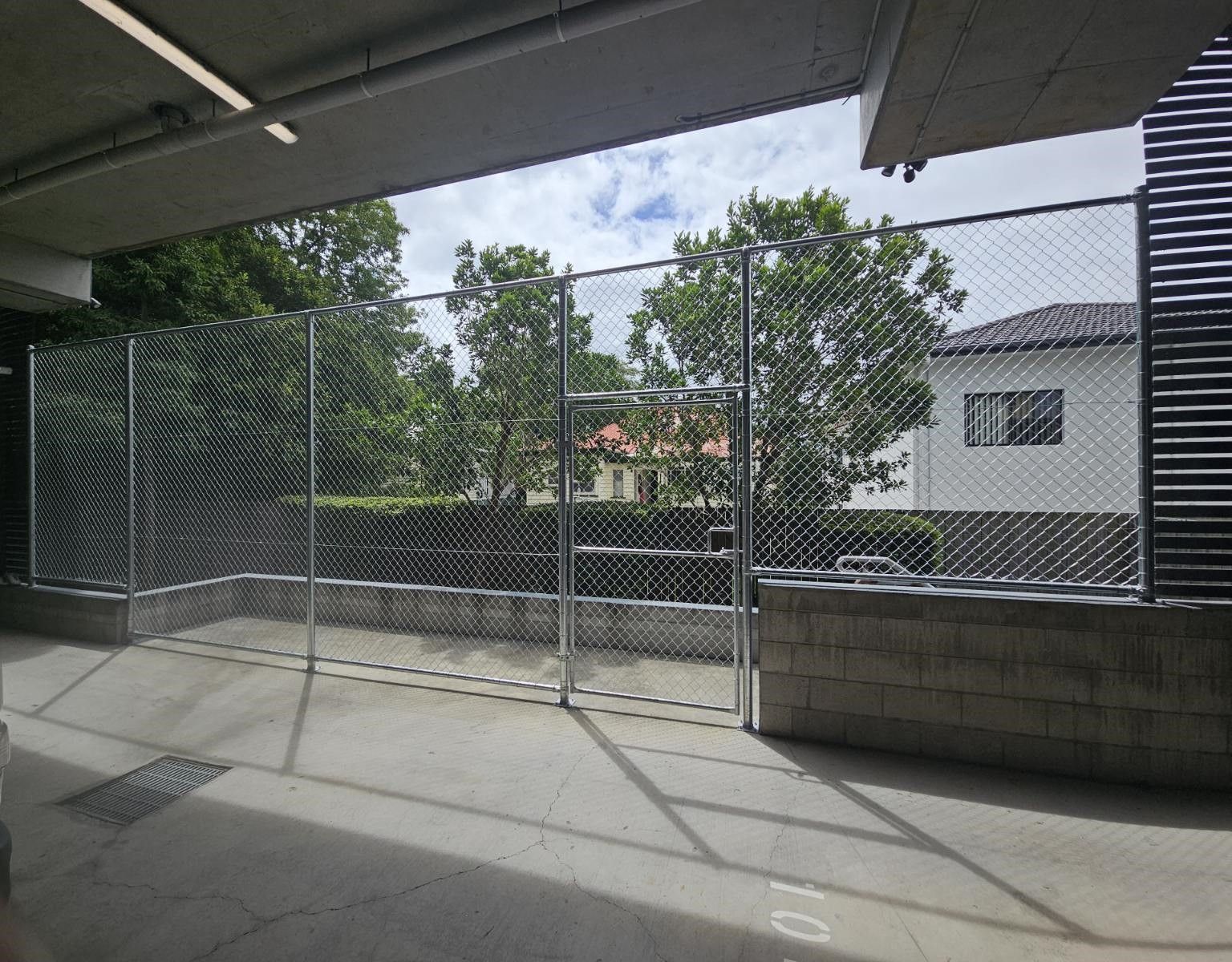 A chain-link fence barrier separates a concrete parking garage area from a residential house and trees in the background.