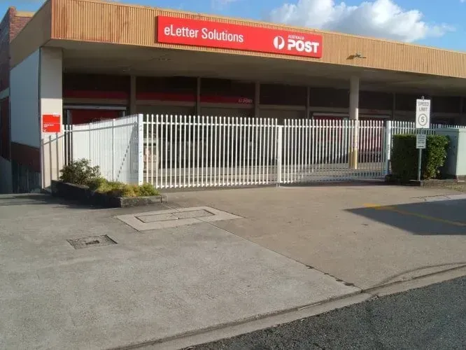 A concrete driveway leads to an eLetter Solutions Australia Post facility behind a white metal fence on a sunny day.