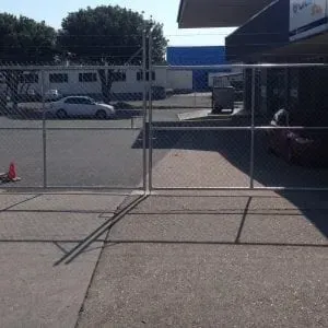 A chain-link fence divides a parking area with a white car in the background from a paved foreground under bright sun.