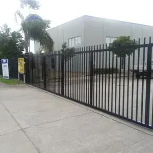 A tall black metal security gate bars the entrance to a building complex on a paved lot with trees.