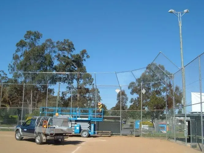 A blue utility truck and a blue scissor lift are parked on a dirt baseball field near a chain-link fence under a clear sky.