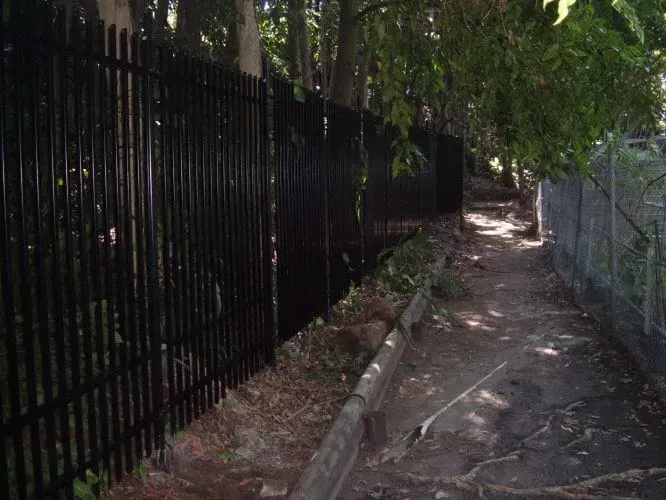 A narrow, shaded dirt path runs between a tall, black metal security fence on the left and a chain-link fence on the right.