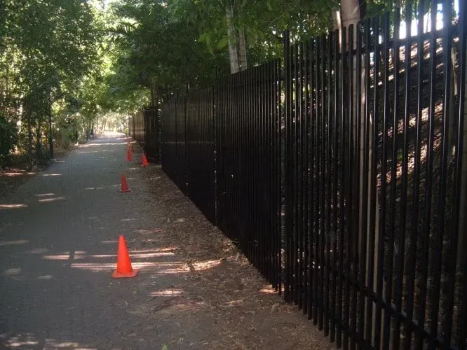 A paved path lined by a tall black metal fence with orange traffic cones placed along the edge.