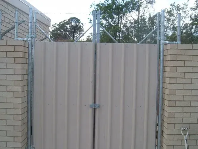 A tan metal double gate set between brick pillars, topped with metal framing and security wire.