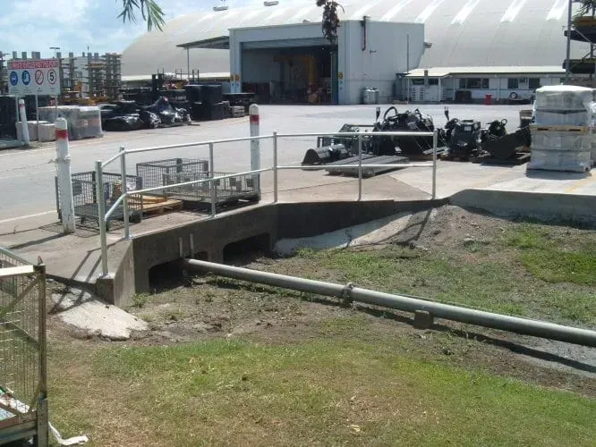 A concrete drainage culvert with a metal railing sits next to a grassy area in an industrial yard with warehouse buildings.